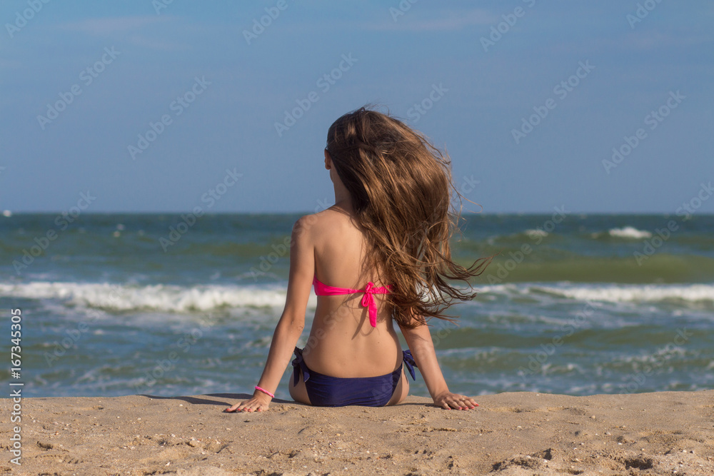 Beautiful young woman in a swimsuit with long hair sits on the beach near the sea. Hot summer day Beautiful young woman in a swimsuit with long hair sits on the beach near the sea. Hot summer day