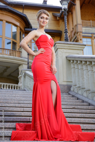 fashion outdoor photo of sexy beautiful woman in luxurious red dress posing on stairs in villa
