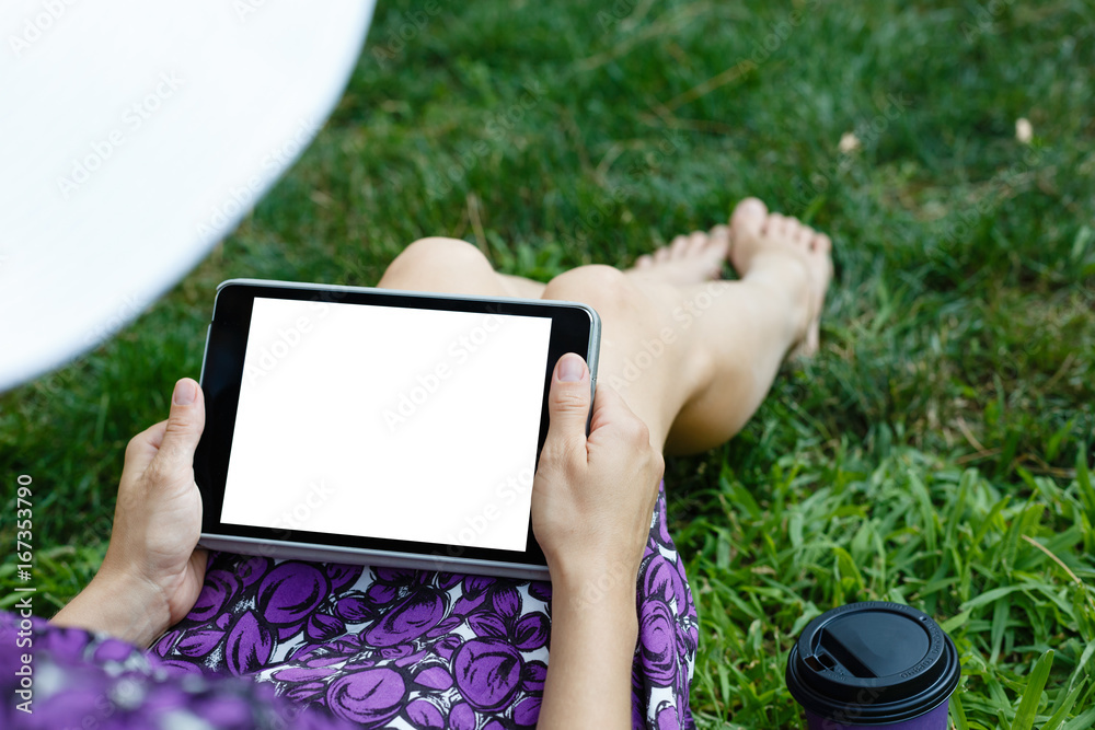 Anonymous woman in white hat and dress relaxing on green grass with tablet. Rear view. Crop shot with vertical orientation screen template.