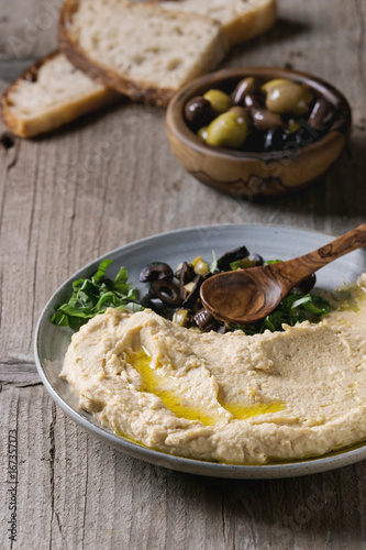 Foto Homemade traditional spread hummus with chopping olives and herbs on blue plate, served with bread, olives variety, olive oil and wood spoon over old wooden background