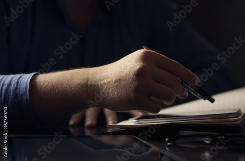 Close-up of a man sitting in a dark-colored workplace and working the pen on the phone and a diary © Bogdan