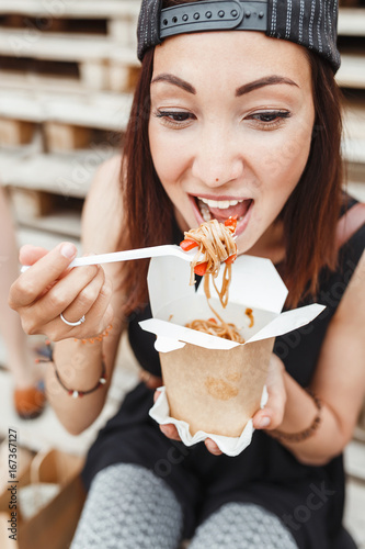 Young Woman Eating Asian wok Noodle Cuisine from Take away Box at the urban street background