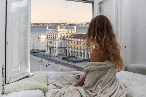 Woman sitting on the bed in the hotel room and looking at view from window at Lisbon city streets and Comercio Square