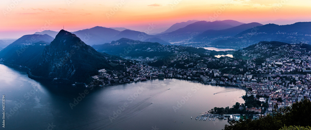 Aerial view of the lake Lugano surrounded by mountains and evening city Lugano on during dramatic sunset, Switzerland, Alps. Travel