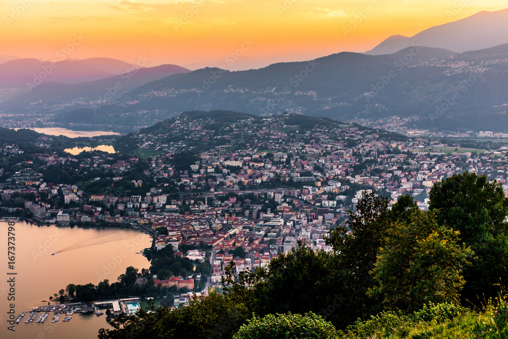 Aerial view of the lake Lugano surrounded by mountains and evening city Lugano on during dramatic sunset, Switzerland, Alps. Travel Aerial view of the lake Lugano surrounded by mountains and evening city Lugano on during dramatic sunset, Switzerland, Alps. Travel