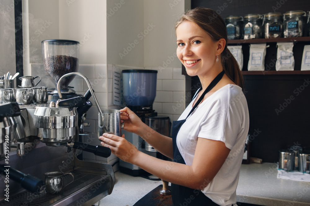 Portrait of happy beautiful young Caucasian smiling woman barista holding milk jug for making coffee using coffee machine. Small local business with organic hot drinks products. Toned with filters. Portrait of happy beautiful young Caucasian smiling woman barista holding milk jug for making coffee using coffee machine. Small local business with organic hot drinks products. Toned with filters.