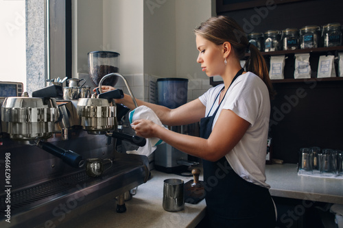 Portrait of young Caucasian woman barista wiping cleaning milk tip of professional espresso coffee machine. Small local business with hot drinks products. Busy life of local small business.