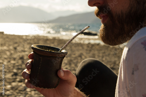 Bearded man drinking mate near a lake.