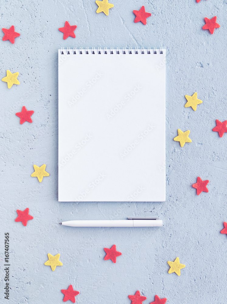 A notebook and a white pen are on a blue concrete background with red and yellow stars. There is an empty space for your text. Vertical image A notebook and a white pen are on a blue concrete background with red and yellow stars. There is an empty space for your text. Vertical image
