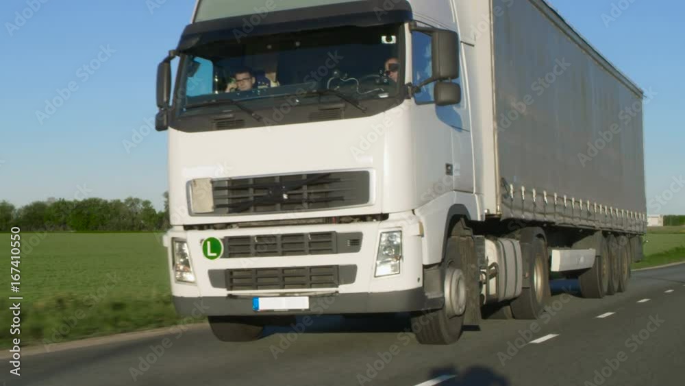 Front-View Camera Follows Semi Truck with Cargo Trailer Driving on a Highway. He's Speeding Through Industrial Warehouse Area while Sun Shines and no other Vehicles are on the Road. Front-View Camera Follows Semi Truck with Cargo Trailer Driving on a Highway. He's Speeding Through Industrial Warehouse Area while Sun Shines and no other Vehicles are on the Road.