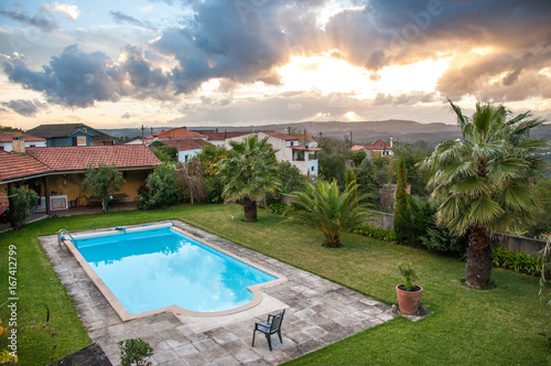 View on a luxury villa and a bright blue pool on a lawn. Shot during a sunset with a dramatic cloudy sky and golden rays shining through the clouds. Shot in Portugal, Villa Nova de Poiares. photo