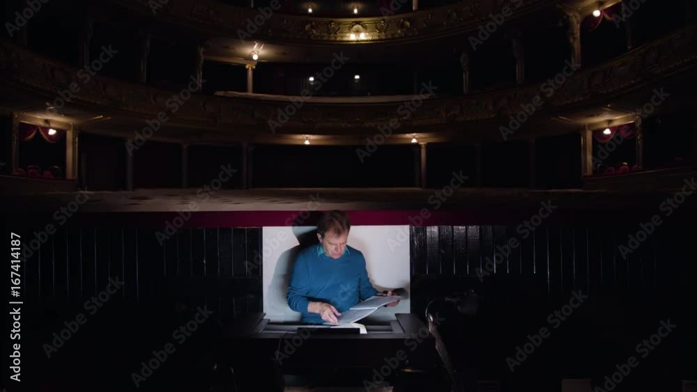 Mid aged conductor alone in dark orchestra pit studies sheet music and tweets the tune. Camera moves away from him. Indoor. Medium Mid aged conductor alone in dark orchestra pit studies sheet music and tweets the tune. Camera moves away from him. Indoor. Medium