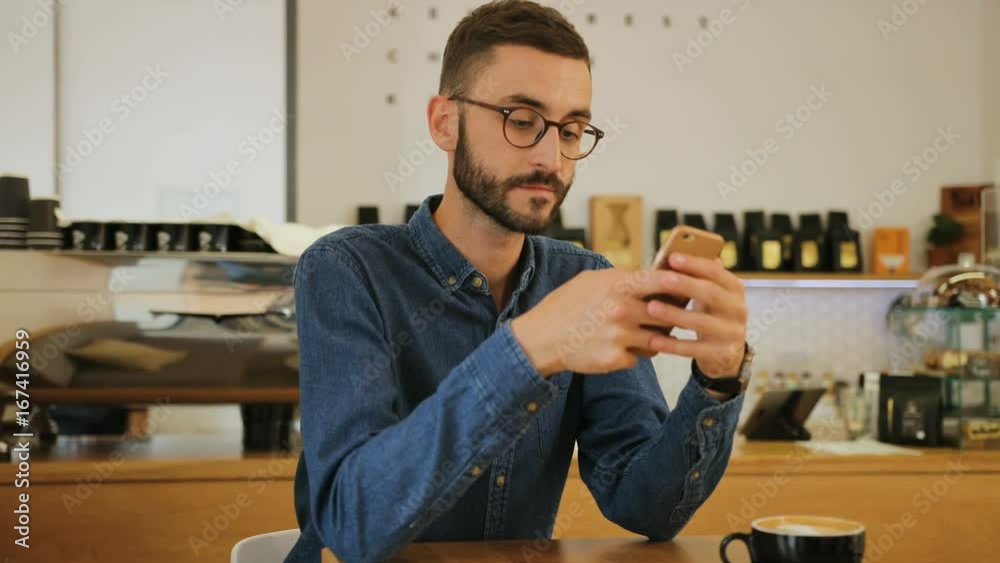 Young attractive man in the glasses using smart phone while sitting at the table in the cafe. Portrait shot.