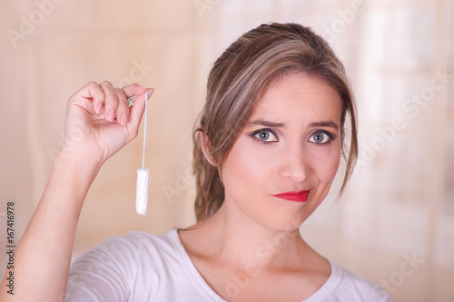 Young beautiful woman with a bored face, holding a menstruation cotton tampon in her hand, in a blurred background