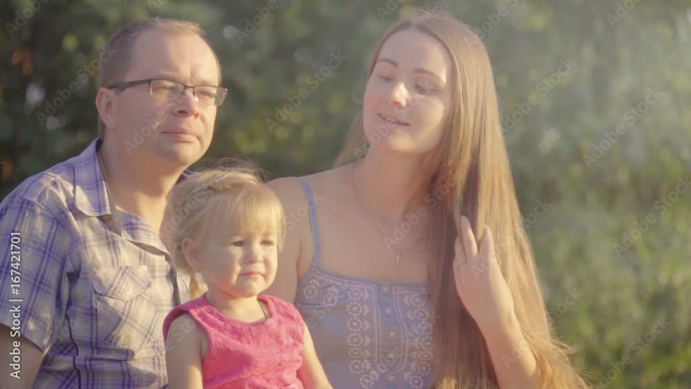 Happy family: Mother, father and little daughter are sitting on the grass in the summer park at sunset. Slow motion Happy family: Mother, father and little daughter are sitting on the grass in the summer park at sunset. Slow motion