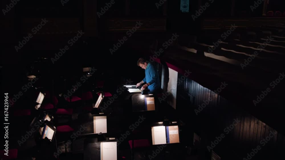 Mid aged conductor alone in dark orchestra pit studies sheet music and tweets the tune. Camera moves from right to left. Indoor. Wide angle Mid aged conductor alone in dark orchestra pit studies sheet music and tweets the tune. Camera moves from right to left. Indoor. Wide angle