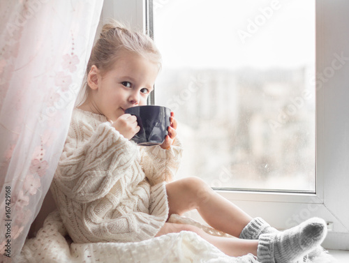 A little girl in the white knitted sweater sits on a window and drinks from a cup