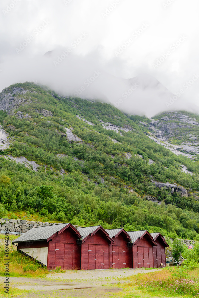 Five red wooden boathouses with dramatic mountainside behind. Rainclouds over the mountain. Location Eidfjord in Norway.