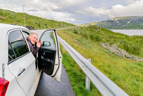 Happy passenger smiling and waving to camera from open door on small car in mountainous landscape. Location Hardangervidda in Norway. © imfotograf