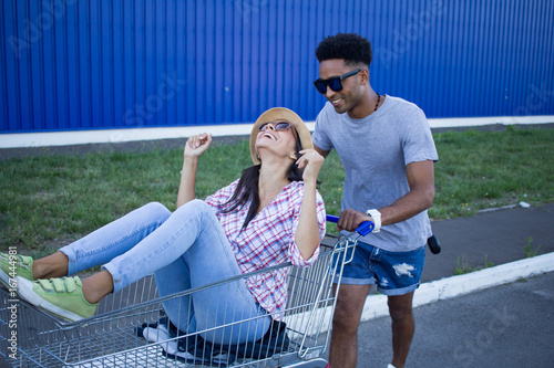 Happy young couple riding on trolley  on empty mall parking , hipster friend have good time during shopping, couple in love riding on shopping cart  photo