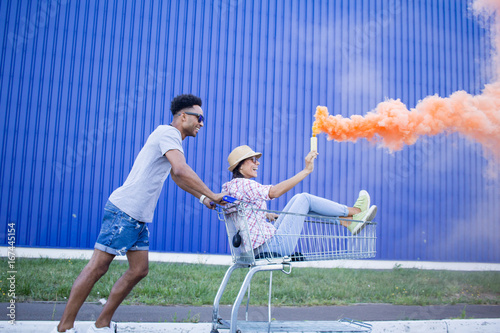 Young beautiful couple in love, boyfriend and girlfriend ride on shopping cart with orange smoke bomb, hipsters having fun in city mall   photo