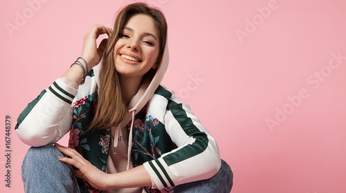 Portrait of a fashionable beautiful laughing girl in a sports jacket print and jeans sitting on a pink background