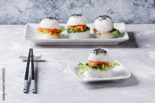 Fotografie Mini rice sushi burgers with smoked salmon, green salad and sauces, black sesame served on white square plate with chopsticks over gray concrete background