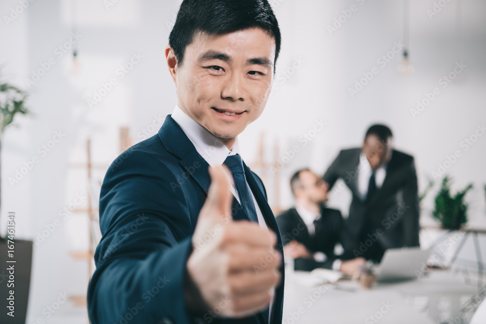 asian businessman showing thumb up with blurred colleagues on background asian businessman showing thumb up with blurred colleagues on background