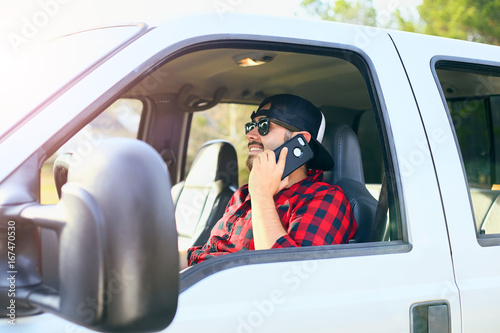 Handsome happy men with a beard smiling in the pickup car truck. Driver talking on mobile cell phone, making phone call. Attractive male driving big vehicle in hat and checkered shirt.