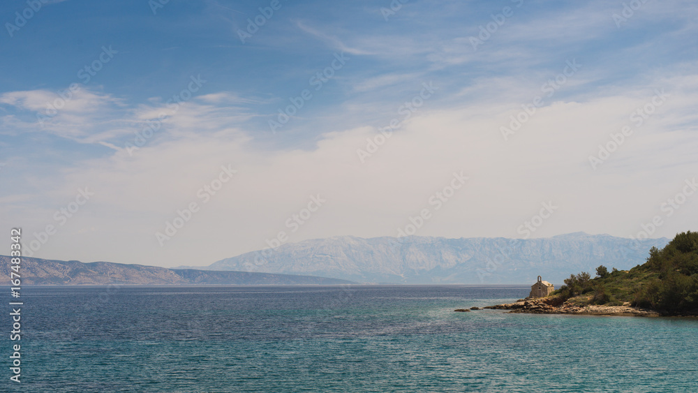 Landscape or panorama of sea horizon with small island with chapel on side, mountains in background, radiant blue sky with cloudy haze. Hvar Island, Brac and Makarska, Croatia.