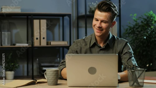 Happy young business man in khaki shirt working on the laptop in the modern office at the evening time, finishing his work and proud of resultats. photo