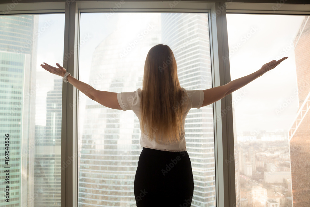 Confident businesswoman spreading hands standing at office window, enjoying big city, successful entrepreneur celebrating business success with arms open wide, feeling powerful inspired, rear view