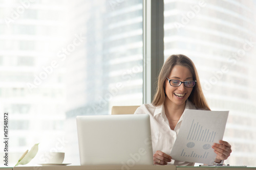 Happy businesswoman holding financial report with rising infographic stats sitting at workplace with laptop, excited by company success, business development, good result and corporate profit growth