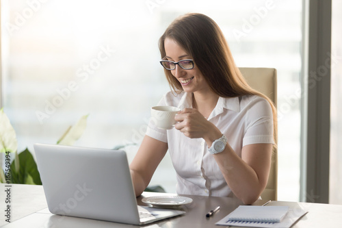 Smiling attractive businesswoman drinking hot caffeine-free coffee in the morning at workplace, happy employee enjoys tasty tea during break at work desk, happy woman holds cup with invigorant drink photo