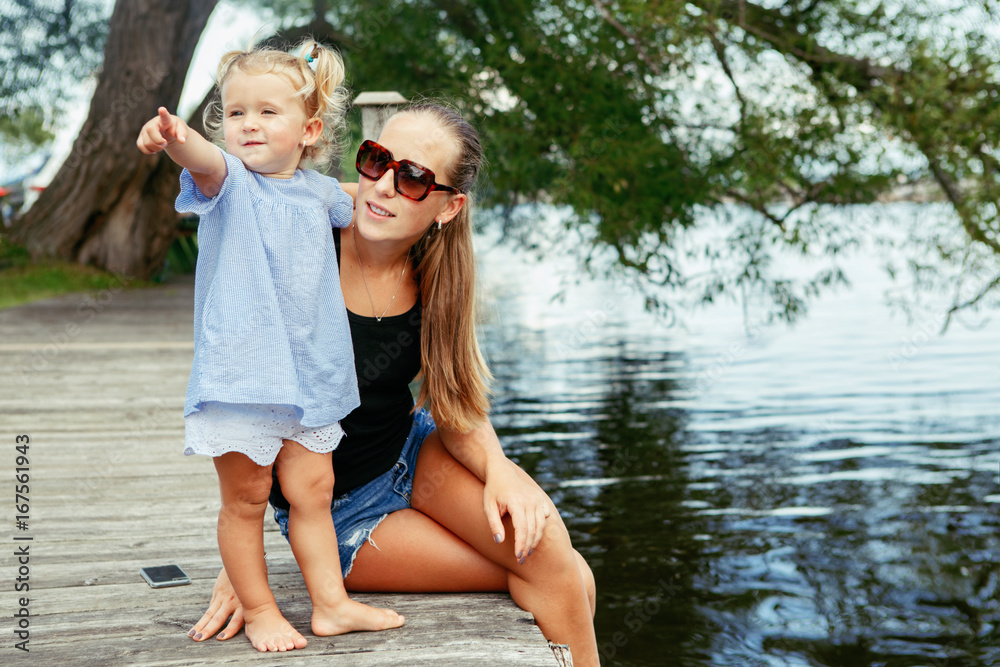 Fototapeta premium Group portrait of happy white Caucasian mother and daughter child having fun outside. Mom and child girl playing, hugging in park on pier by water lake. Candid authentic real lifestyle.