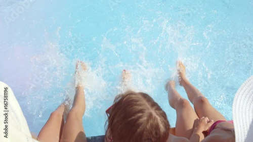 SLOW MOTION CLOSE UP LENS FLARE Three unrecognizable cheerful girls sitting on pool edge, splashing water with their feet on hot sunny day. Three playful young women on summer vacation splashing water