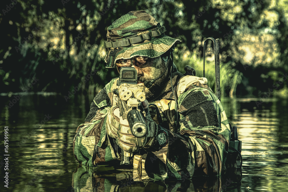 Bearded soldier in action during river raid in the jungle pointing rifle to camera. He is waist deep in the water and mud and ready to meet enemy, survive and fight in agressive hostile environment