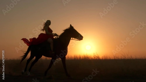Young girl in red dress riding black horse against the sun. Rider with her stallion trotting across a field at sunset. Long gown blowing in the wind. Horseback riding in slow motion.
