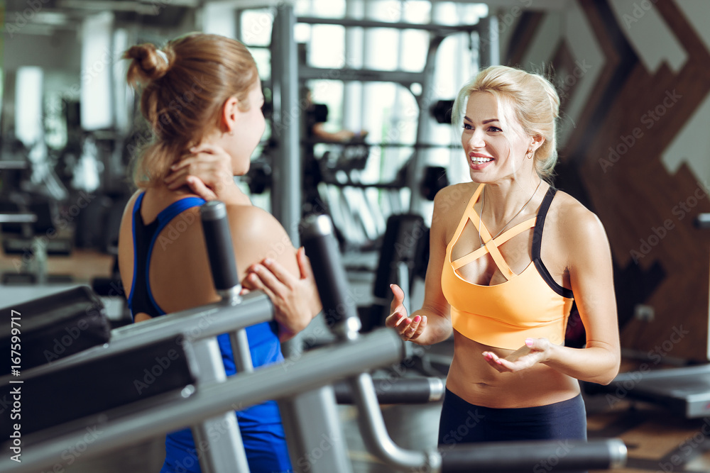 Two athletic blond women talking in gym. Girl communicates with trainer