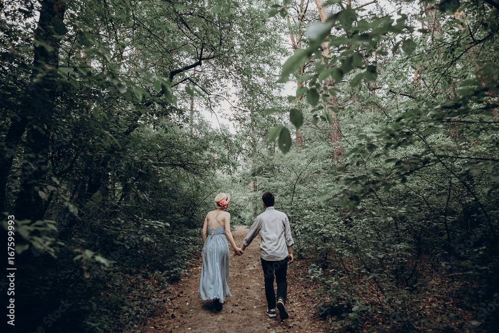 stylish hipster bride and groom walking in green summer forest. happy couple in love, modern outfit, relaxing at park. girl in dress and straw hat with peony. rustic wedding concept.