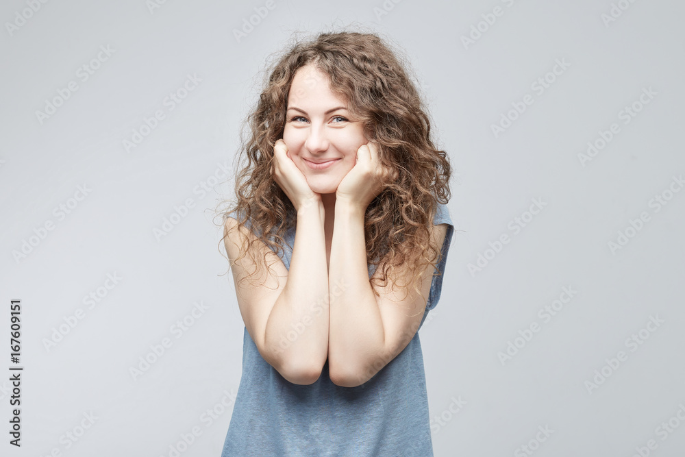 Portrait of contented curly-haired European woman looking with her blue warm eyes and exited smile having sencere and gentle look holding hands undeer chin. People, beauty, emotions concept.