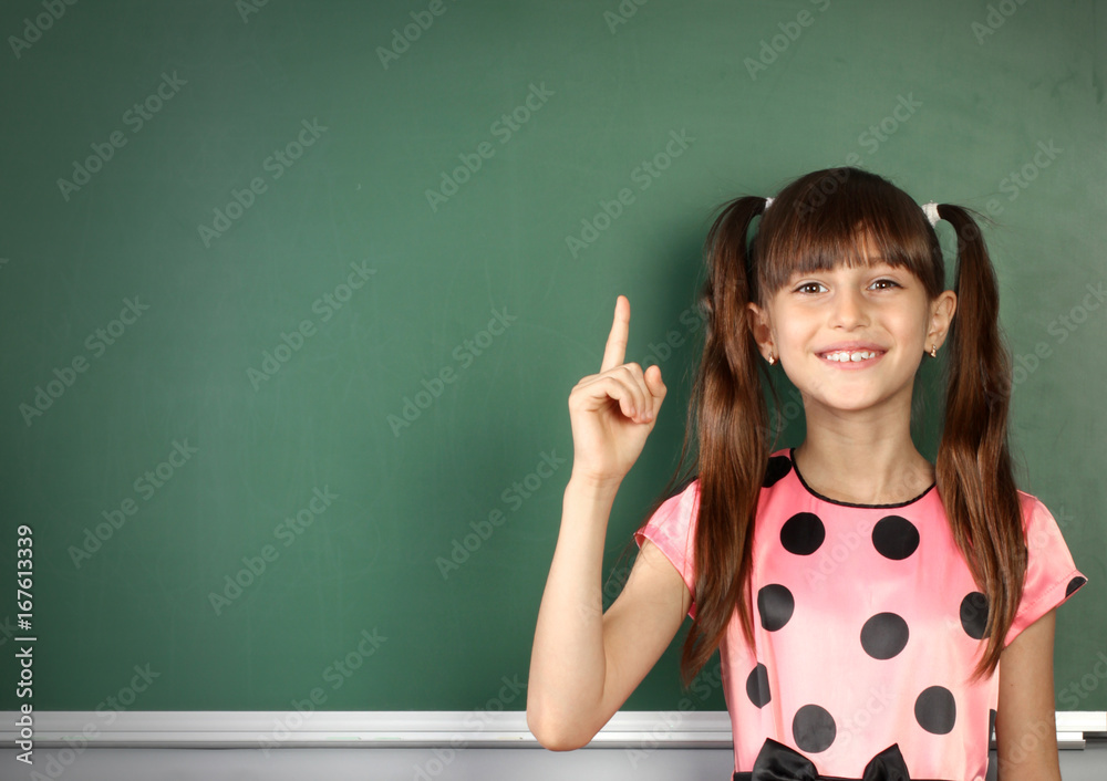 Smiling child girl show with a finger empty school blackboard, copy space