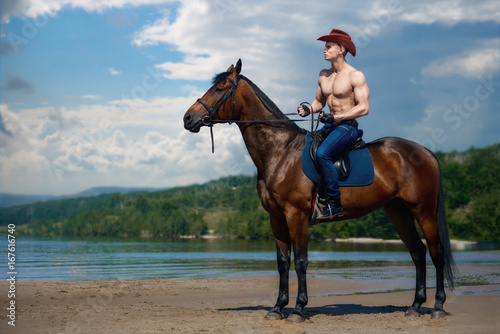 Macho man handsome cowboy riding on a horse on the background of sky and water.