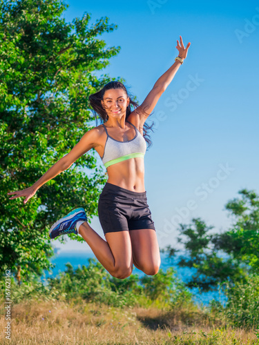 Young Active Sporty Successful Woman Jumping on the high hill above sea or ocean and looking at camera. Beautiful girl in sportswear. Health concept.