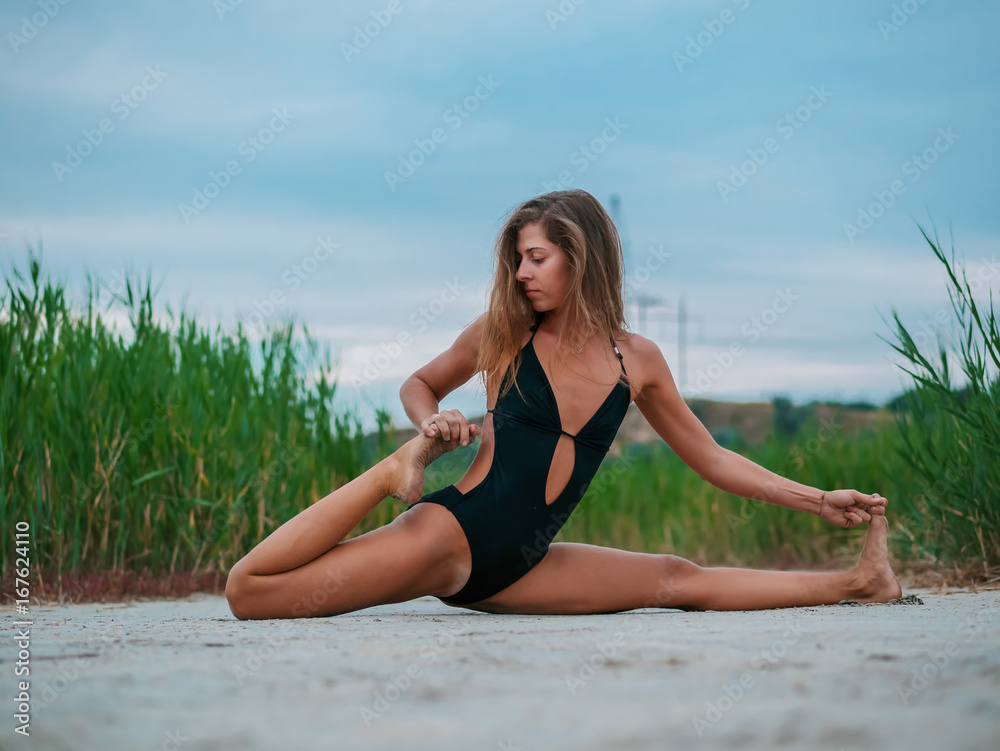 Morning exercise. Twine. Woman stretching hip, hamstring muscles and groin area. Fit fitness athlete girl exercising sports stretches in black swimsuit at sunset near reeds. Morning exercise. Twine. Woman stretching hip, hamstring muscles and groin area. Fit fitness athlete girl exercising sports stretches in black swimsuit at sunset near reeds.