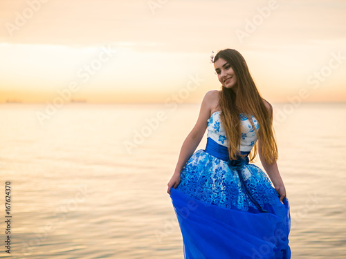 The young beautiful girl dances in a blue evening or ball dress on the pier close to sea. photo