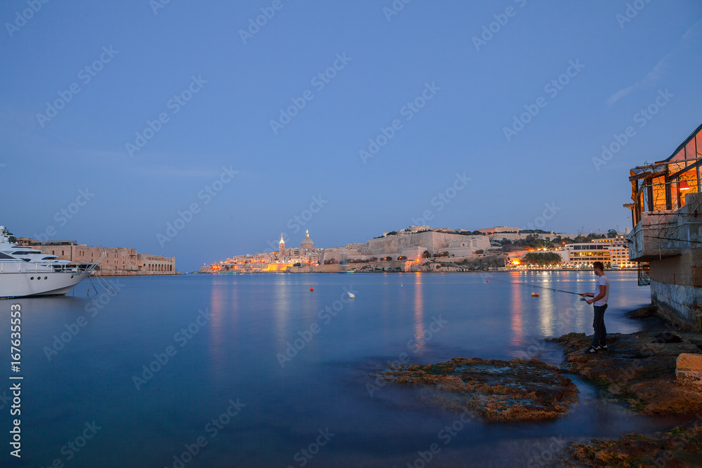Summer night view of Valetta profile over sea. Long exposure. Illuminated architecture. Guy fishing on the shore.