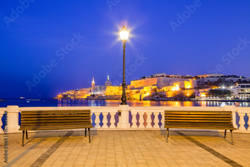 Summer night view of the embankment with benches and lantern and Valetta profile over sea at the background. Long exposure. Illuminated architecture.