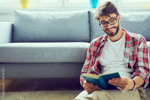 Portrait of joyful young man reading book while sitting on floor in his living room