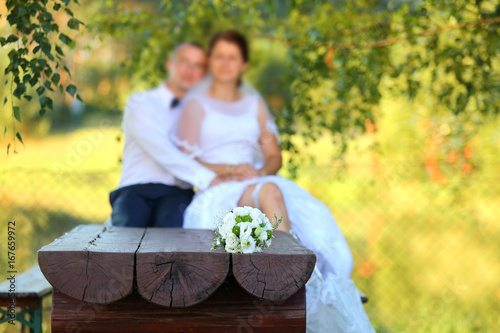 Bukiet ślubny na drewnianej ławie z młodą parą w tle.